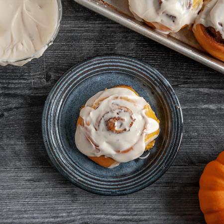 top down view of pumpkin cinnamon roll iced on a white plate with pumpkin and fall setting on table